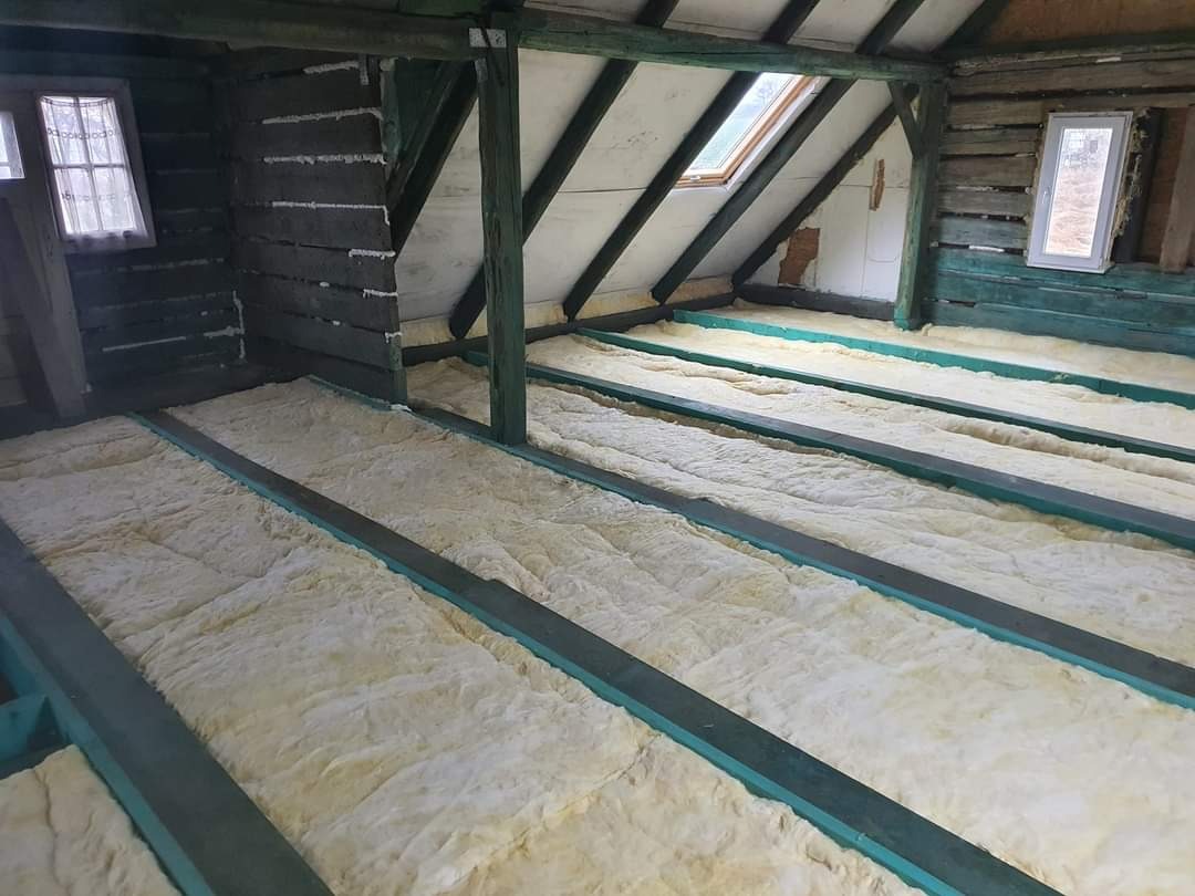 Wnętrze poddasza w trakcie ocieplania: wełna mineralna ułożona między drewnianymi belkami, widoczne okno dachowe i fragmenty drewnianej konstrukcji ścian.