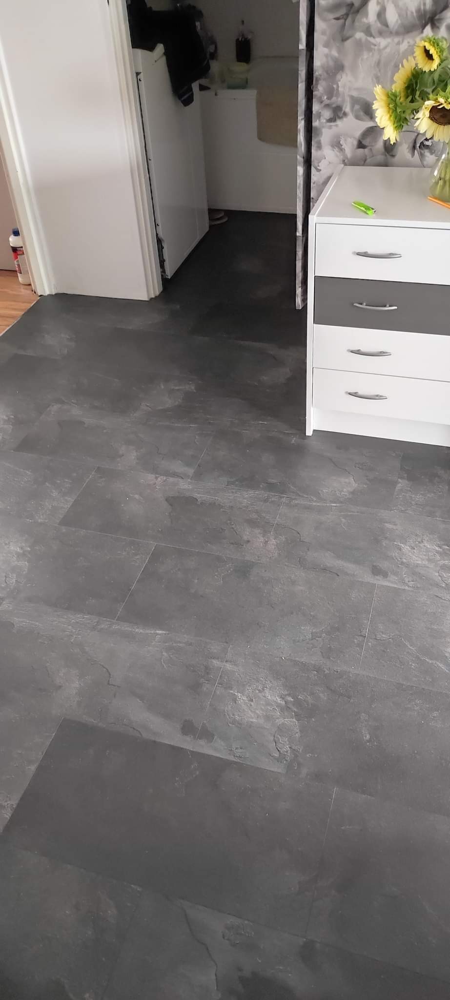 Nowoczesna, ciemnoszara podłoga z płytek imitujących kamień, ułożona w pomieszczeniu z białymi ścianami i szafką z kwiatami w tle.