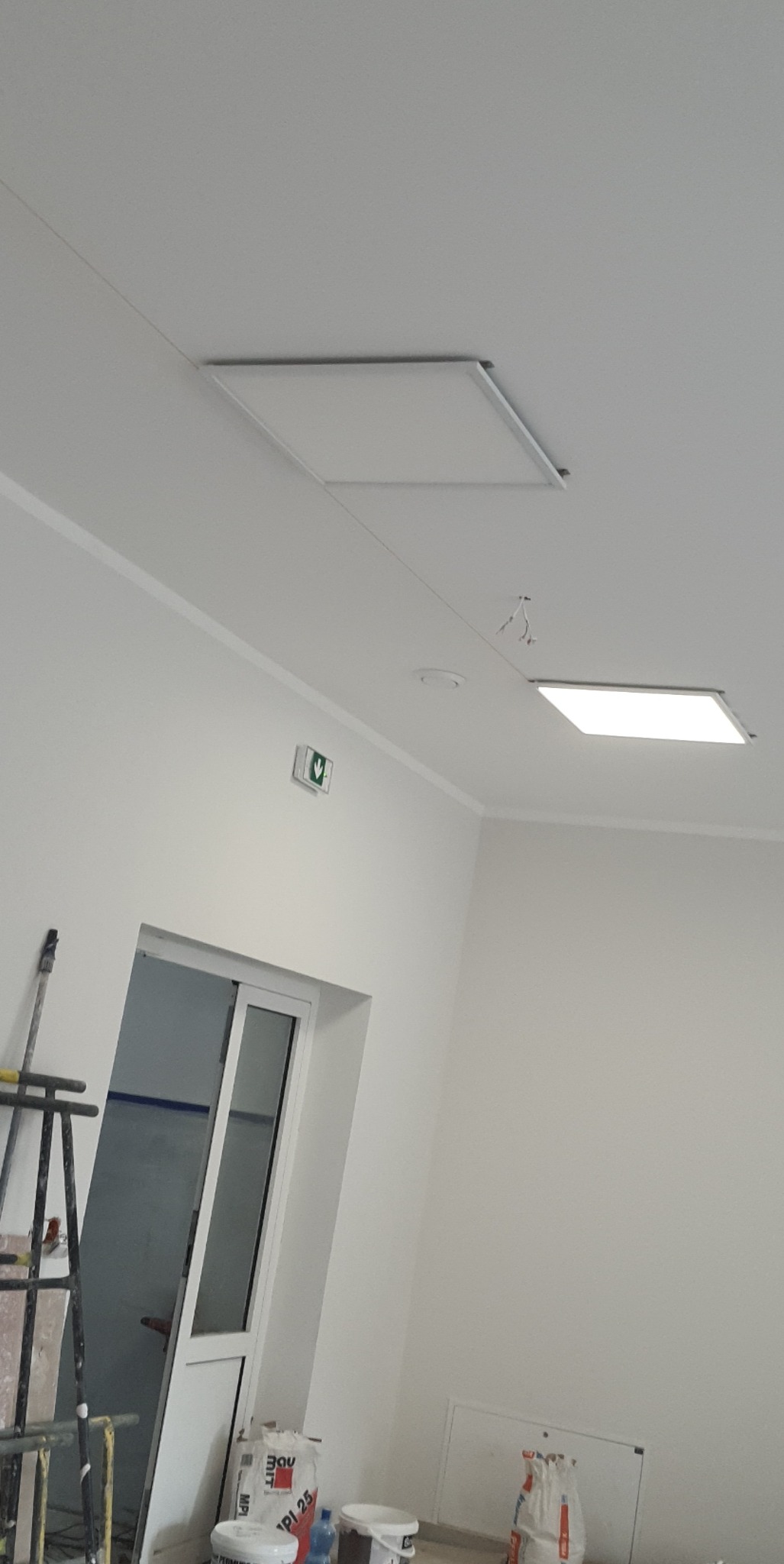 Wnętrze po odświeżeniu: pomalowane na biało ściany z listwami, nowe panele sufitowe, widoczny fragment drabiny malarskiej i worki z zaprawą przy drzwiach.