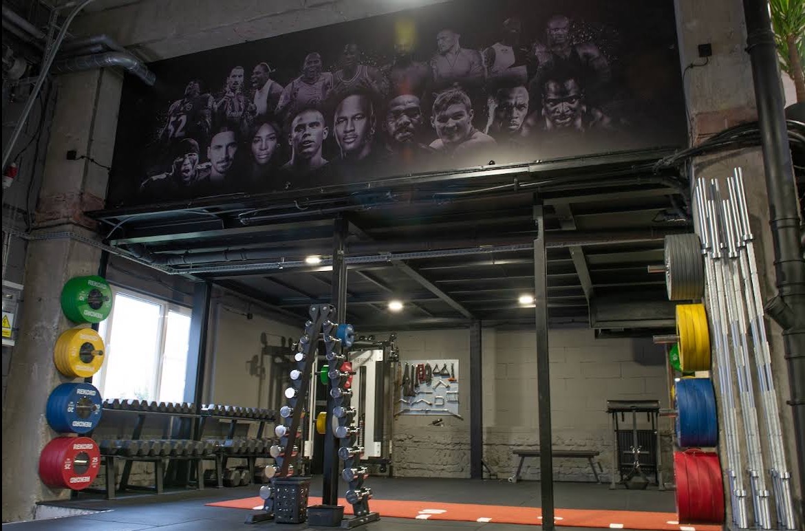 Profesjonalna siłownia z ciężarami, sztangami i motywacyjnym muralem z wizerunkami sportowców na ścianie. Nowoczesny design i sprzęt do ćwiczeń.
