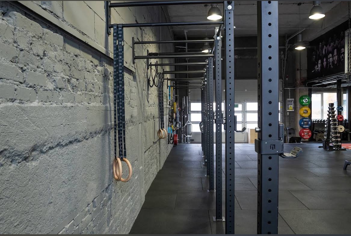Nowoczesna siłownia z szarymi ścianami, wyposażona w drabinki gimnastyczne, kółka i stojaki na obciążenia. Widok w głąb sali, oświetlenie sufitowe.