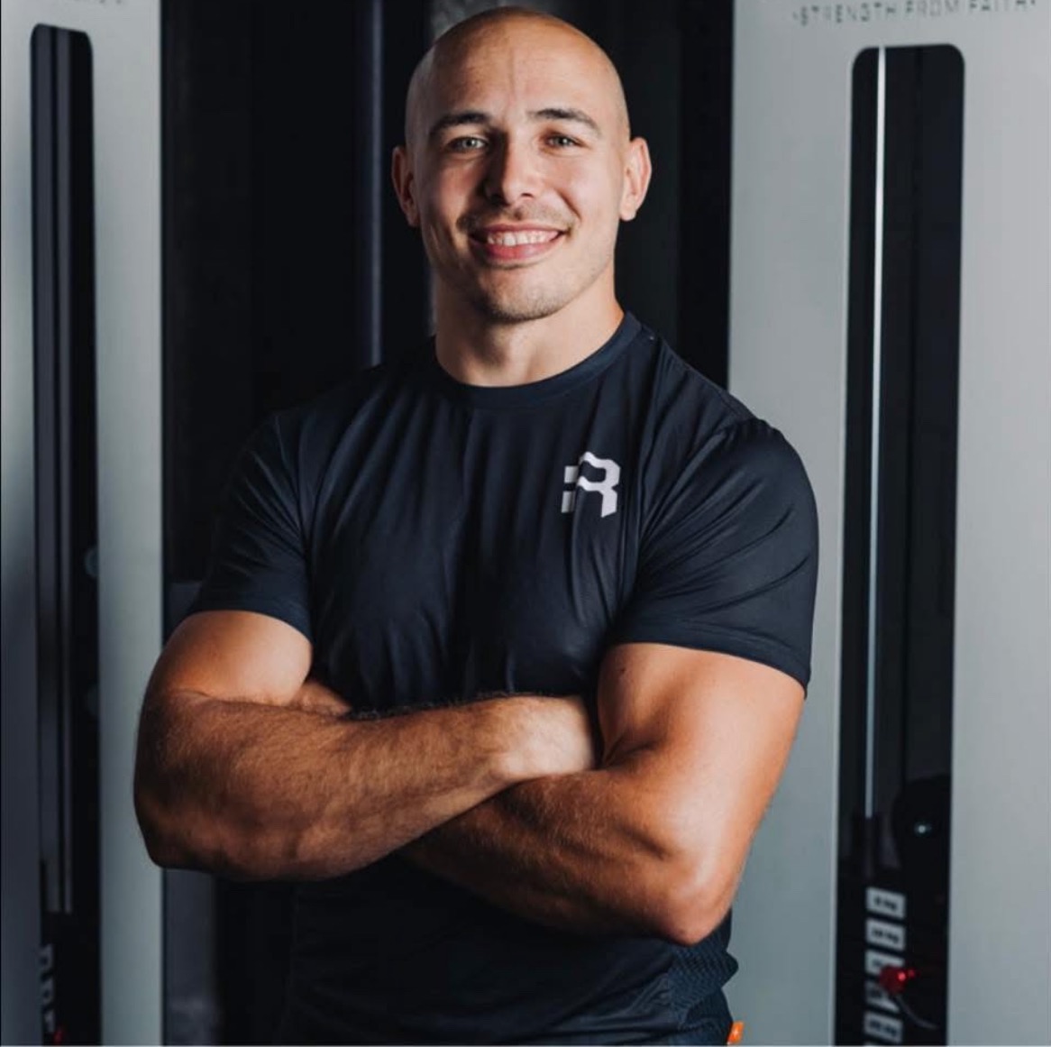 Uśmiechnięty mężczyzna we Wrocławiu, z muskularnymi ramionami skrzyżowanymi na tle sprzętu do ćwiczeń. Czarna koszulka z logo. Profesjonalny wygląd.