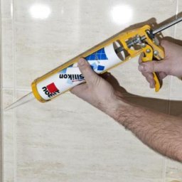 Hydraulik - Nakładanie silikonu z pistoletu na fugę między płytkami ceramicznymi w łazience, widoczne ręce i fragment ramienia osoby wykonującej prace.