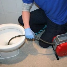 Hydraulik - Hydraulik w niebieskich rękawicach udrażnia toaletę za pomocą elektrycznej sprężyny do rur, czerwona obudowa urządzenia stoi na podłodze.