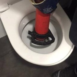 Hydraulik - Udrażnianie toalety przy użyciu czerwonego przepychacza z czarną gumową podstawą, widoczne dłonie w roboczych rękawicach i fragment spodni osoby wykonującej pracę.