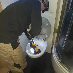 Hydraulik - Hydraulik w roboczych spodniach i rękawicach, z lampą czołową, używa przepychacza z żółtą rączką do udrożnienia muszli klozetowej w łazience z kabiną prysznicową.