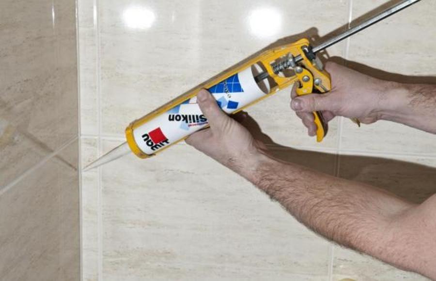 Nakładanie silikonu z pistoletu na fugę między płytkami ceramicznymi w łazience, widoczne ręce i fragment ramienia osoby wykonującej prace.
