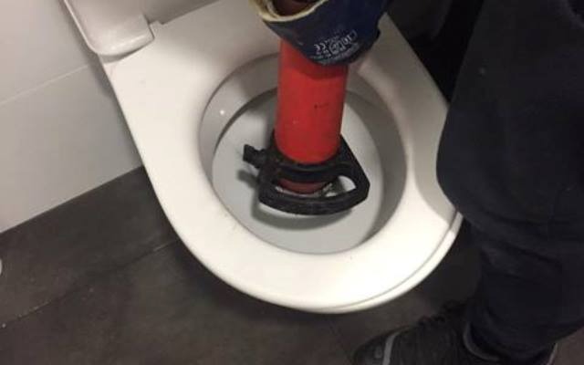 Udrażnianie toalety przy użyciu czerwonego przepychacza z czarną gumową podstawą, widoczne dłonie w roboczych rękawicach i fragment spodni osoby wykonującej pracę.