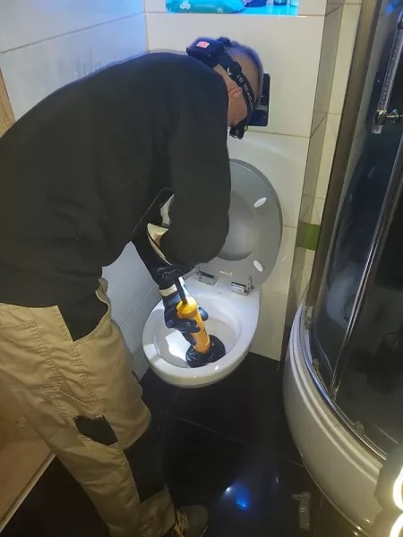 Hydraulik w roboczych spodniach i rękawicach, z lampą czołową, używa przepychacza z żółtą rączką do udrożnienia muszli klozetowej w łazience z kabiną prysznicową.