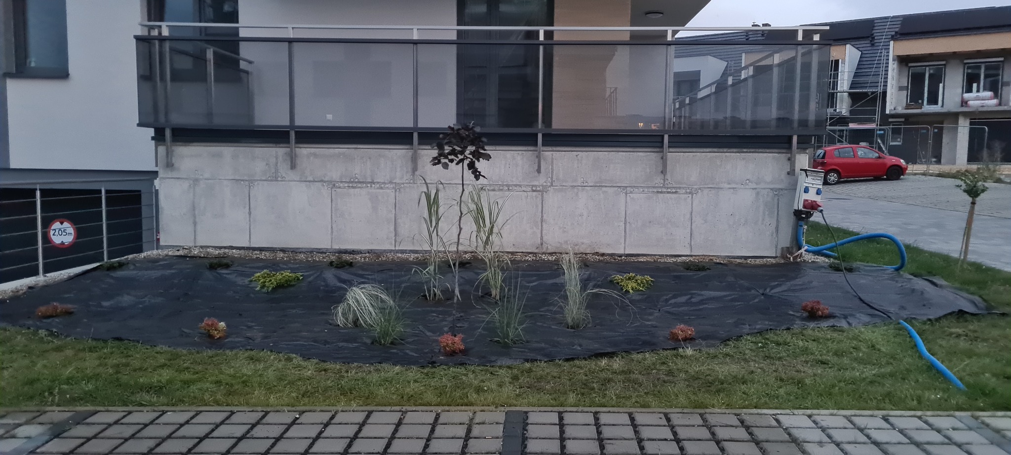 Nowo założony ogród z czarną agrowłókniną, świeżo posadzone rośliny ozdobne, trawy i małe drzewko, w tle betonowy budynek mieszkalny i czerwony samochód.