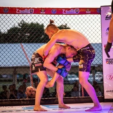 Dwóch zawodników w klatce MMA podczas walki: jeden zawodnik próbuje obalić drugiego, trzymając go w pasie, widoczne logo Octagon na spodenkach jednego z zawodników.