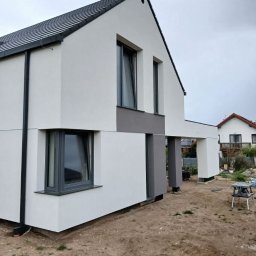 Mariusz Filek - Nowoczesny dom jednorodzinny z biało-szarą elewacją, ciemną dachówką i szarą stolarką okienną, widok z boku na tle pochmurnego nieba.