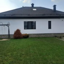 Biały dom jednorodzinny z ciemną dachówką, widoczny z perspektywy trawnika z huśtawką i ozdobnymi krzewami, z elementami wykończenia elewacji imitującymi kamień.