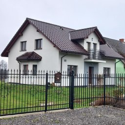 Mariusz Filek - Biały dom jednorodzinny z brązowym dachem i balkonem, otoczony czarnym, kutym ogrodzeniem na lekko pochylonym terenie.