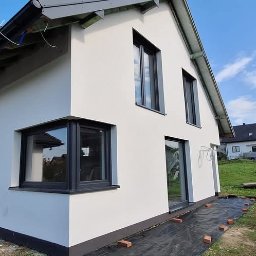 Mariusz Filek - Ocieplona elewacja domu jednorodzinnego z szarymi oknami, widok z zewnątrz na tle błękitnego nieba i innego budynku w oddali.