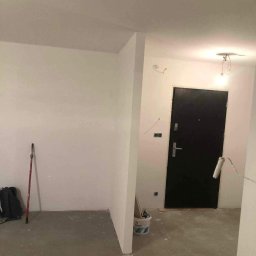 Mariusz Filek - Wnętrze w trakcie malowania: pomalowane na biało ściany, widoczny wałek malarski oparty o czarne drzwi wejściowe, wiadro z farbą gruntującą w rogu pomieszczenia.