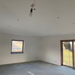 Mariusz Filek - Wnętrze w stanie deweloperskim z gładkimi, białymi ścianami, szarą wylewką podłogową i prowizorycznym oświetleniem w postaci wiszącej żarówki. Widoczne okno z widokiem na zewnątrz i przesuwne drzwi...