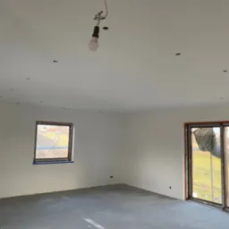 Wnętrze w stanie deweloperskim z gładkimi, białymi ścianami, szarą wylewką podłogową i prowizorycznym oświetleniem w postaci wiszącej żarówki. Widoczne okno z widokiem na zewnątrz i przesuwne drzwi...