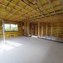 Mariusz Filek - Wnętrze domu w trakcie budowy: widoczne drewniane ramy konstrukcyjne ścian i sufitu, ocieplenie ścian żółtą wełną mineralną Eurovent, okno z białymi szprosami i surowa podłoga.