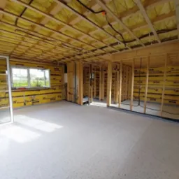 Wnętrze domu w trakcie budowy: widoczne drewniane ramy konstrukcyjne ścian i sufitu, ocieplenie ścian żółtą wełną mineralną Eurovent, okno z białymi szprosami i surowa podłoga.