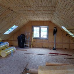 Mariusz Filek - Poddasze w trakcie izolacji: widoczne belki, wełna mineralna na ścianach i poddaszu, okno dachowe, drabina oparta o ścianę, materiały budowlane, poziomica.