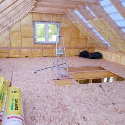 Mariusz Filek - Adaptacja poddasza: widok na ocieplone skośne ściany i sufit z widoczną konstrukcją drewnianą, częściowo ułożoną podłogą z płyt OSB, drabina aluminiowa z butelką wody, rolki wełny mineralnej...