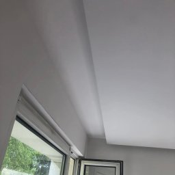 Mariusz Filek - Wnętrze po remoncie: świeżo pomalowany sufit i ściany w jasnym odcieniu, widoczne okno z widokiem na zieleń.