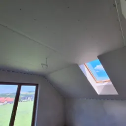 Poddasze w trakcie wykańczania płytami gipsowo-kartonowymi, widoczne okno dachowe z błękitnym niebem i fragment krajobrazu za oknem ściennym.