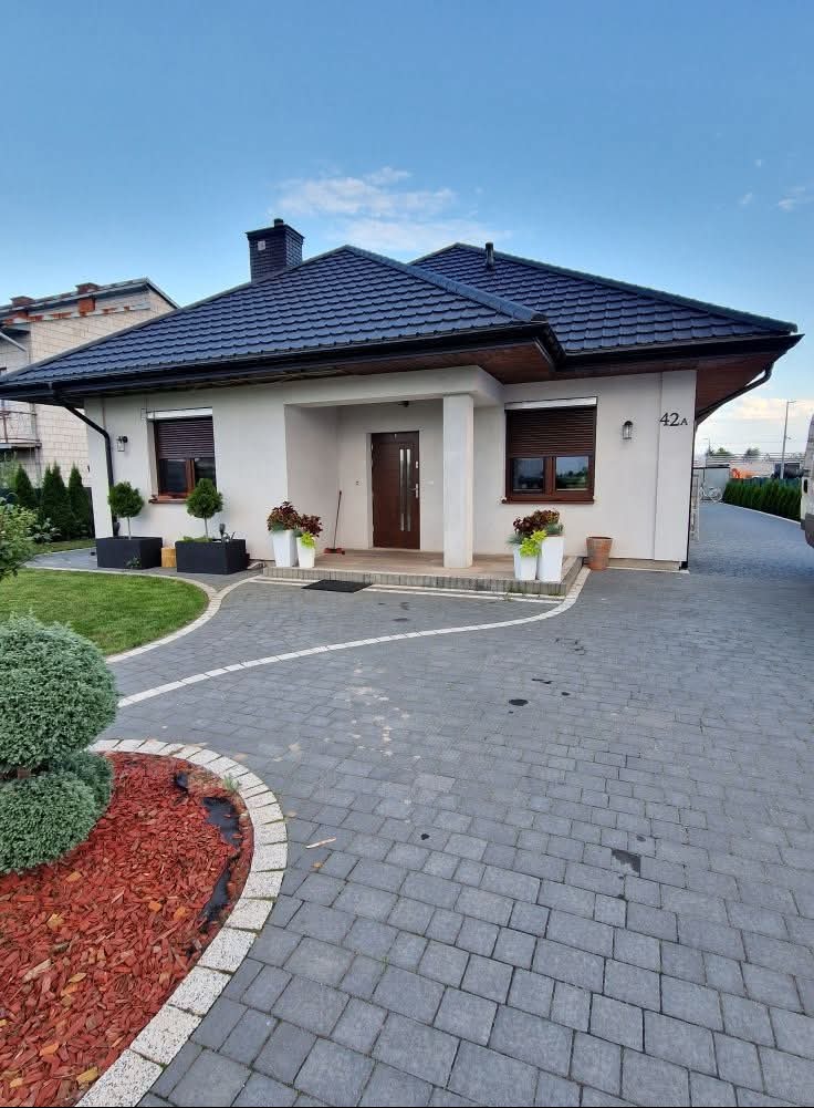 Nowoczesny dom jednorodzinny z ciemną dachówką, jasną elewacją z numerem 42a, drewnianą stolarką okienną i brukowanym podjazdem. Przed wejściem donice z kwiatami.
