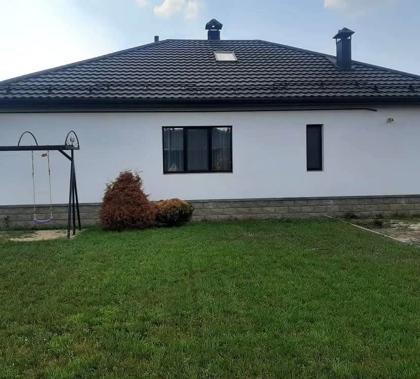 Biały dom jednorodzinny z ciemną dachówką, widoczny z perspektywy trawnika z huśtawką i ozdobnymi krzewami, z elementami wykończenia elewacji imitującymi kamień.