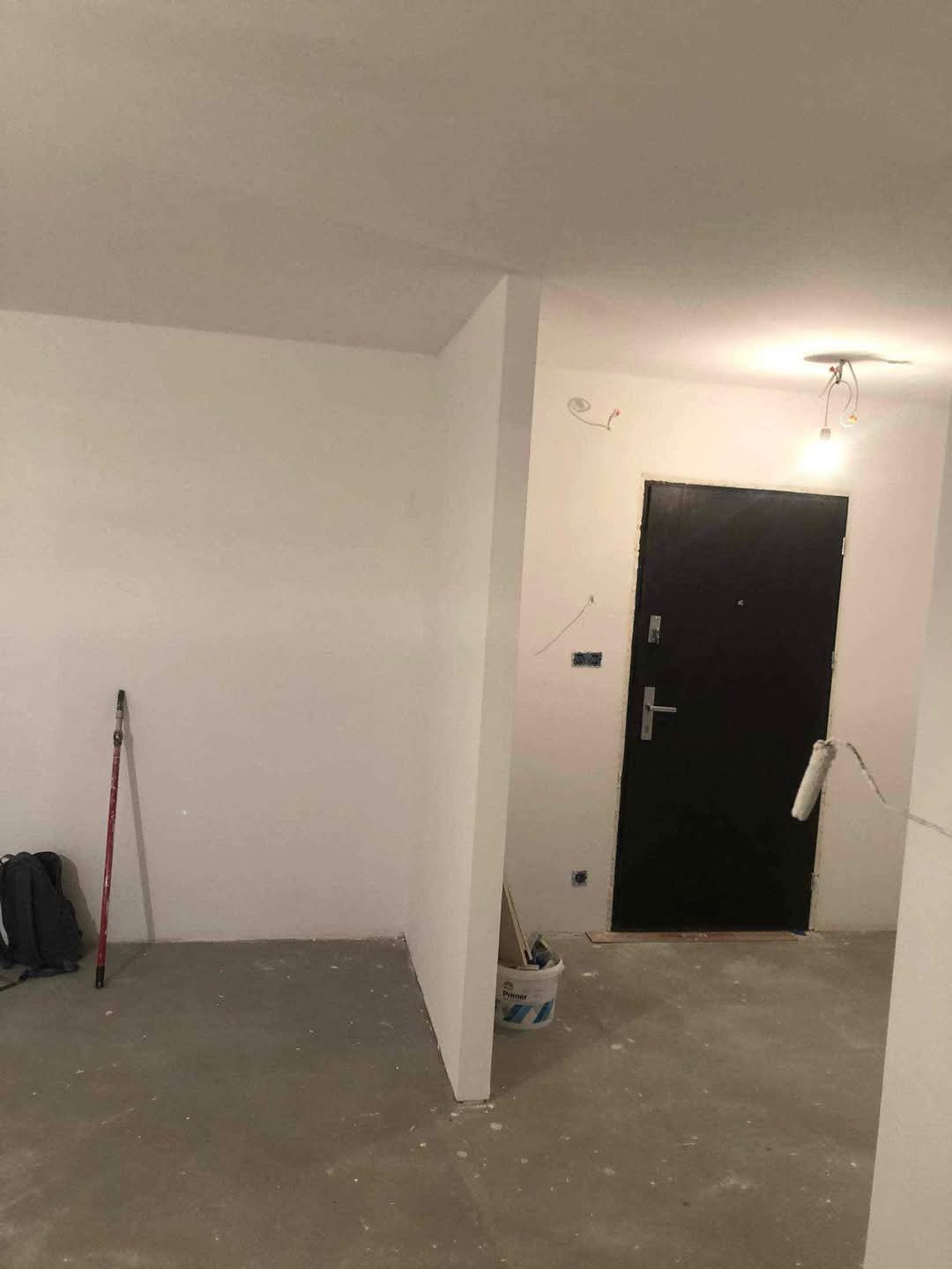 Wnętrze w trakcie malowania: pomalowane na biało ściany, widoczny wałek malarski oparty o czarne drzwi wejściowe, wiadro z farbą gruntującą w rogu pomieszczenia.