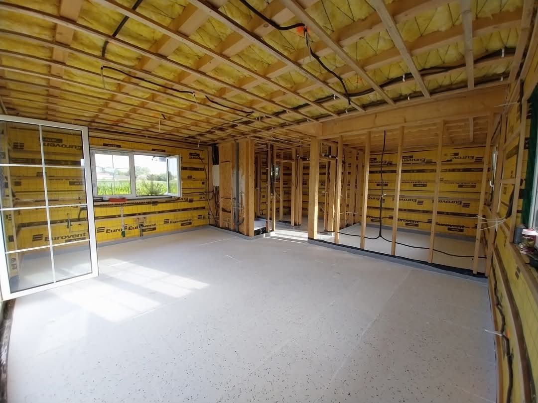 Wnętrze domu w trakcie budowy: widoczne drewniane ramy konstrukcyjne ścian i sufitu, ocieplenie ścian żółtą wełną mineralną Eurovent, okno z białymi szprosami i surowa podłoga.