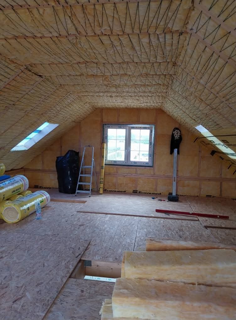 Poddasze w trakcie izolacji: widoczne belki, wełna mineralna na ścianach i poddaszu, okno dachowe, drabina oparta o ścianę, materiały budowlane, poziomica.