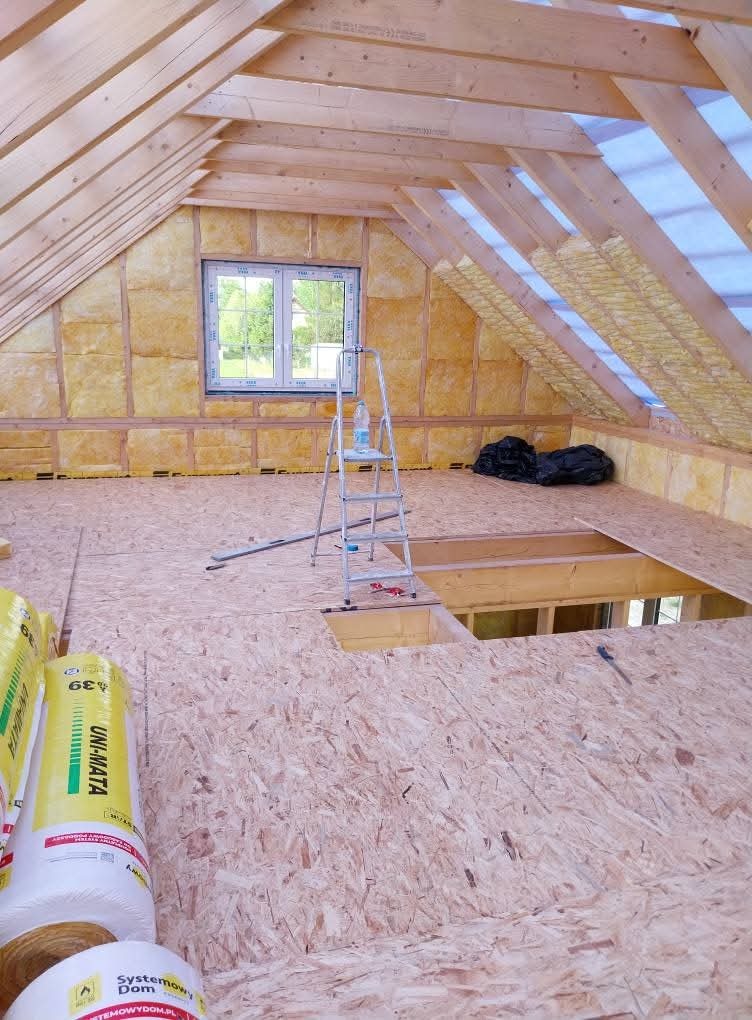 Adaptacja poddasza: widok na ocieplone skośne ściany i sufit z widoczną konstrukcją drewnianą, częściowo ułożoną podłogą z płyt OSB, drabina aluminiowa z butelką wody, rolki wełny mineralnej...