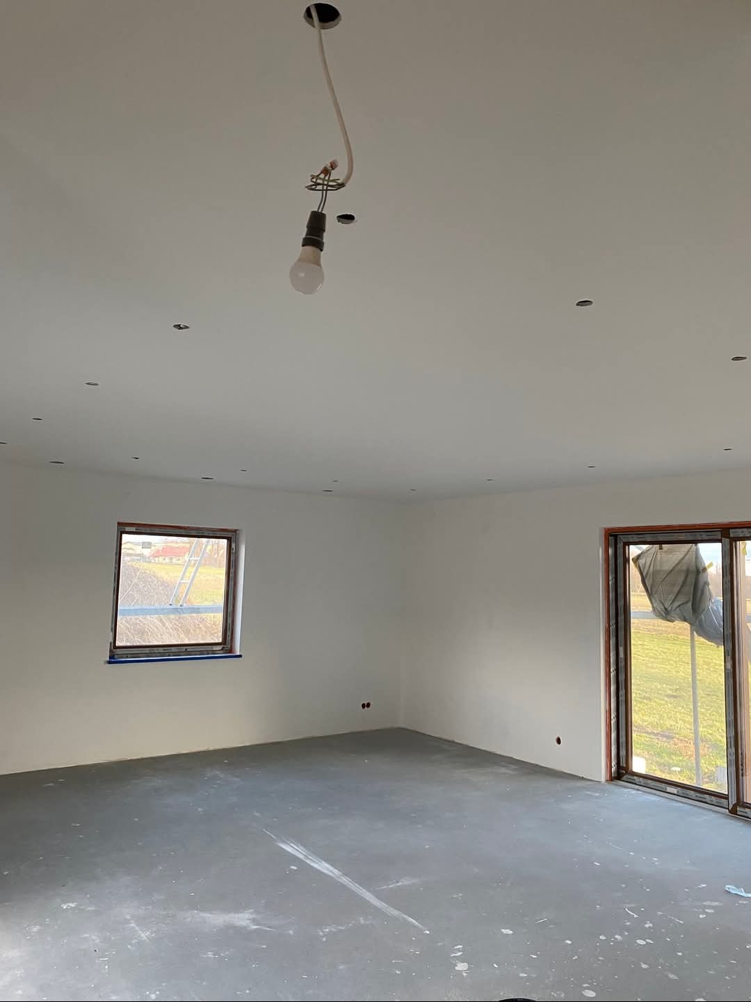 Wnętrze w stanie deweloperskim po szpachlowaniu i malowaniu na biało, widoczne ślady po pracach na podłodze, okno z widokiem na zewnątrz i prowizoryczne oświetlenie.