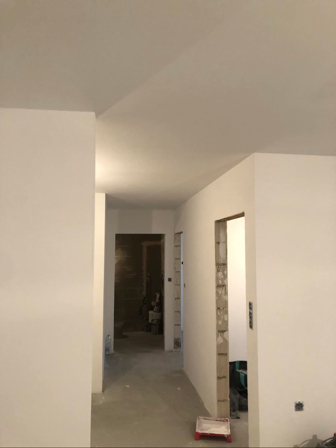 Korytarz w trakcie remontu, widoczne ściany po szpachlowaniu i malowaniu na biało, surowy beton wokół ościeżnic, podłoga przygotowana do dalszych prac wykończeniowych, pojemnik z resztkami farby...