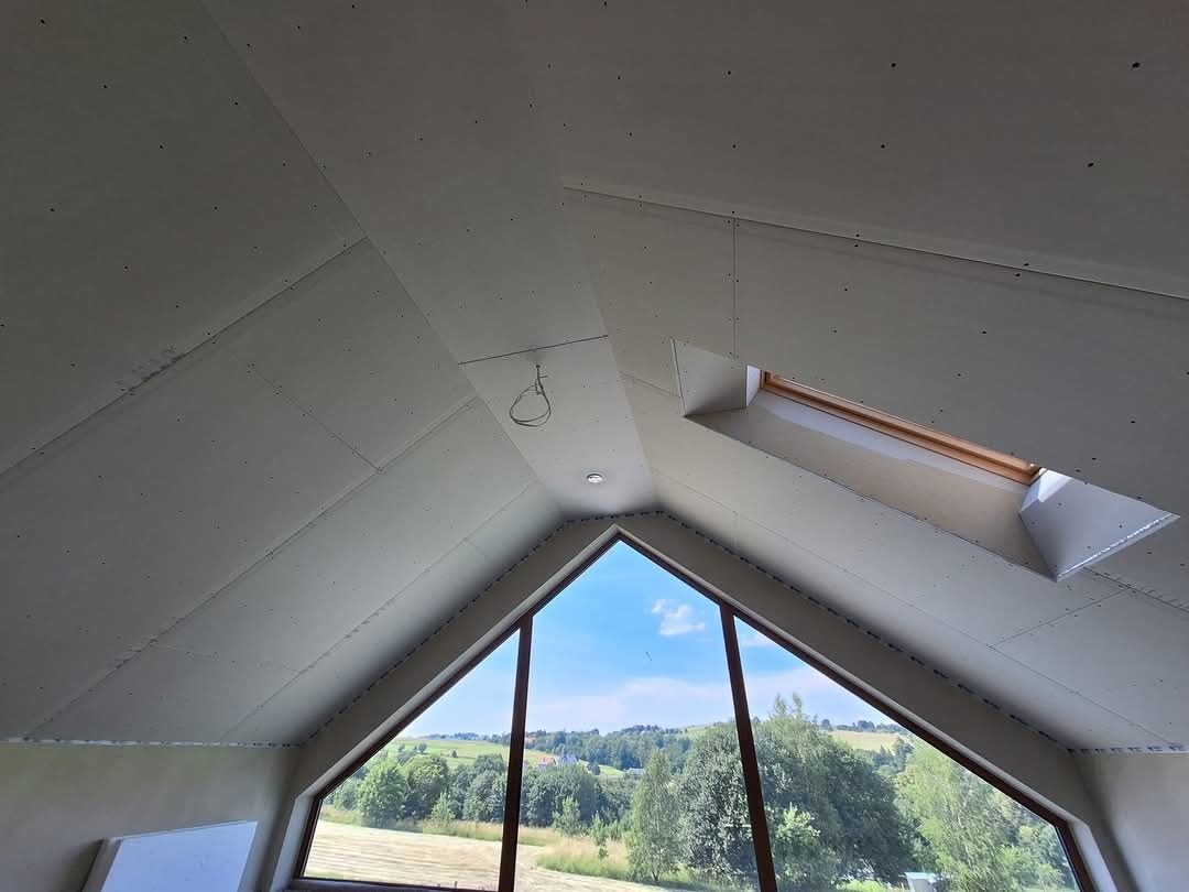 Montaż płyt gipsowo-kartonowych na poddaszu ze skośnym oknem dachowym, widok na zielony krajobraz za oknem trójkątnym.