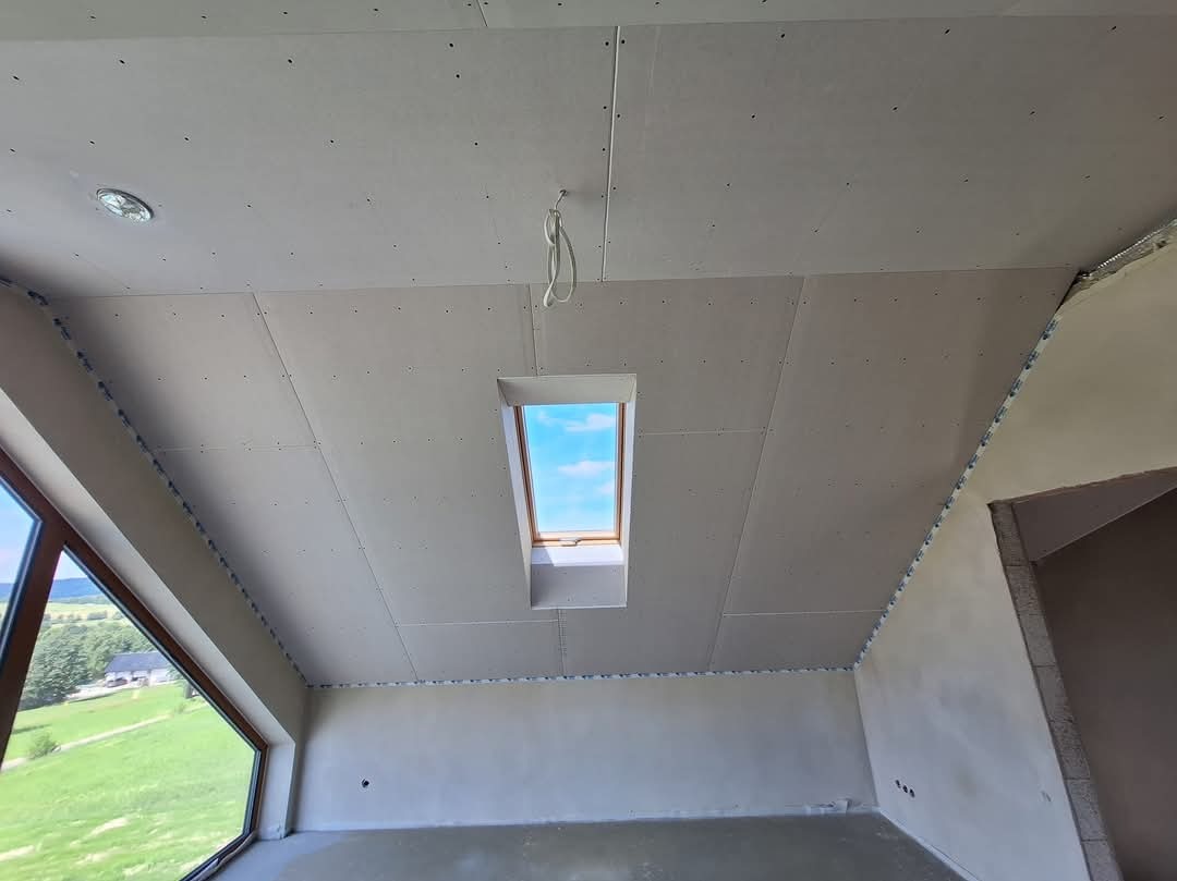 Poddasze w trakcie wykańczania: widok na zamontowane płyty gipsowo-kartonowe na skośnym suficie z oknem dachowym i wystającymi przewodami elektrycznymi, ściany częściowo pokryte gładzią.