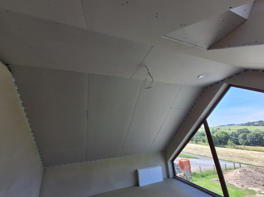 Poddasze z zamontowanymi płytami kartonowo-gipsowymi na skosach dachu i suficie, widoczne łączenia płyt, surowe ściany, okno z widokiem na krajobraz.