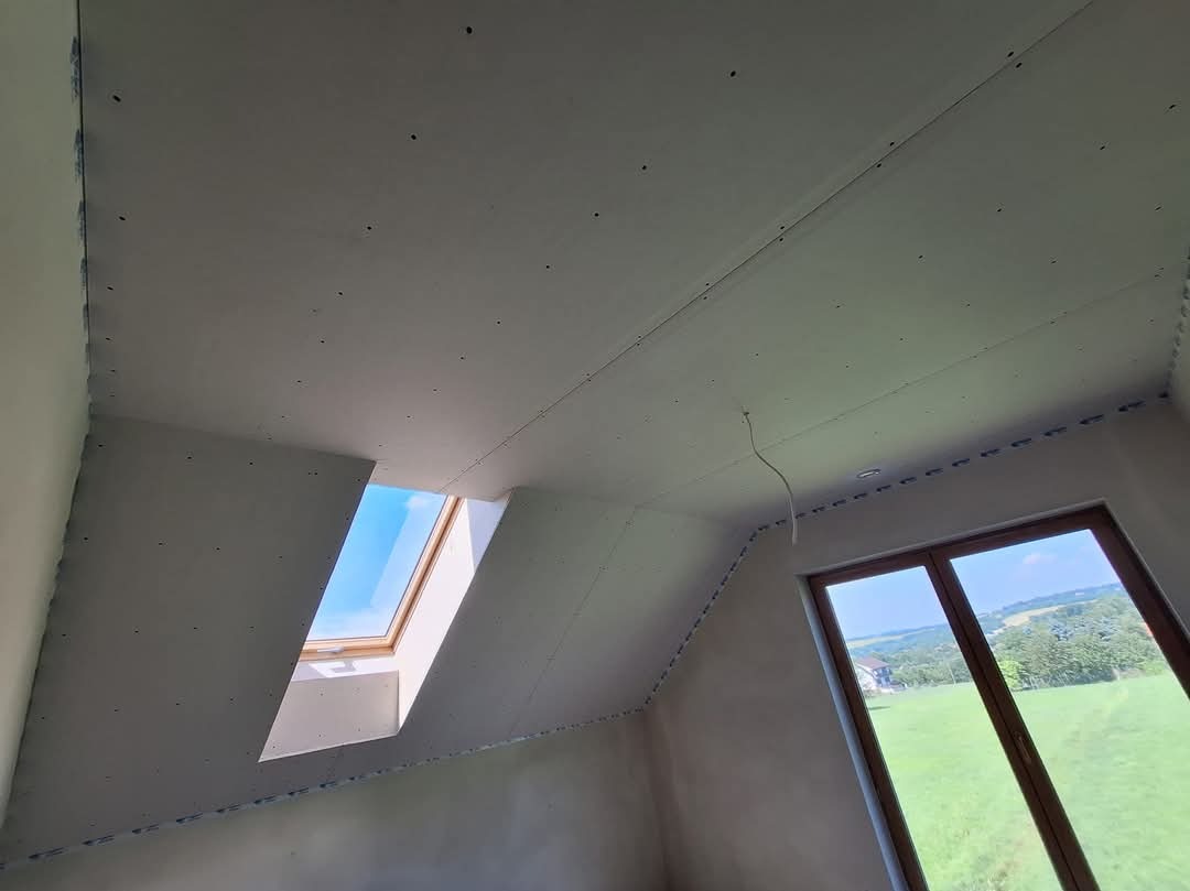 Montaż płyt karton gips na poddaszu ze skośnym dachem, widoczne profile i łączenia, okno dachowe i okno pionowe z widokiem na zieleń.