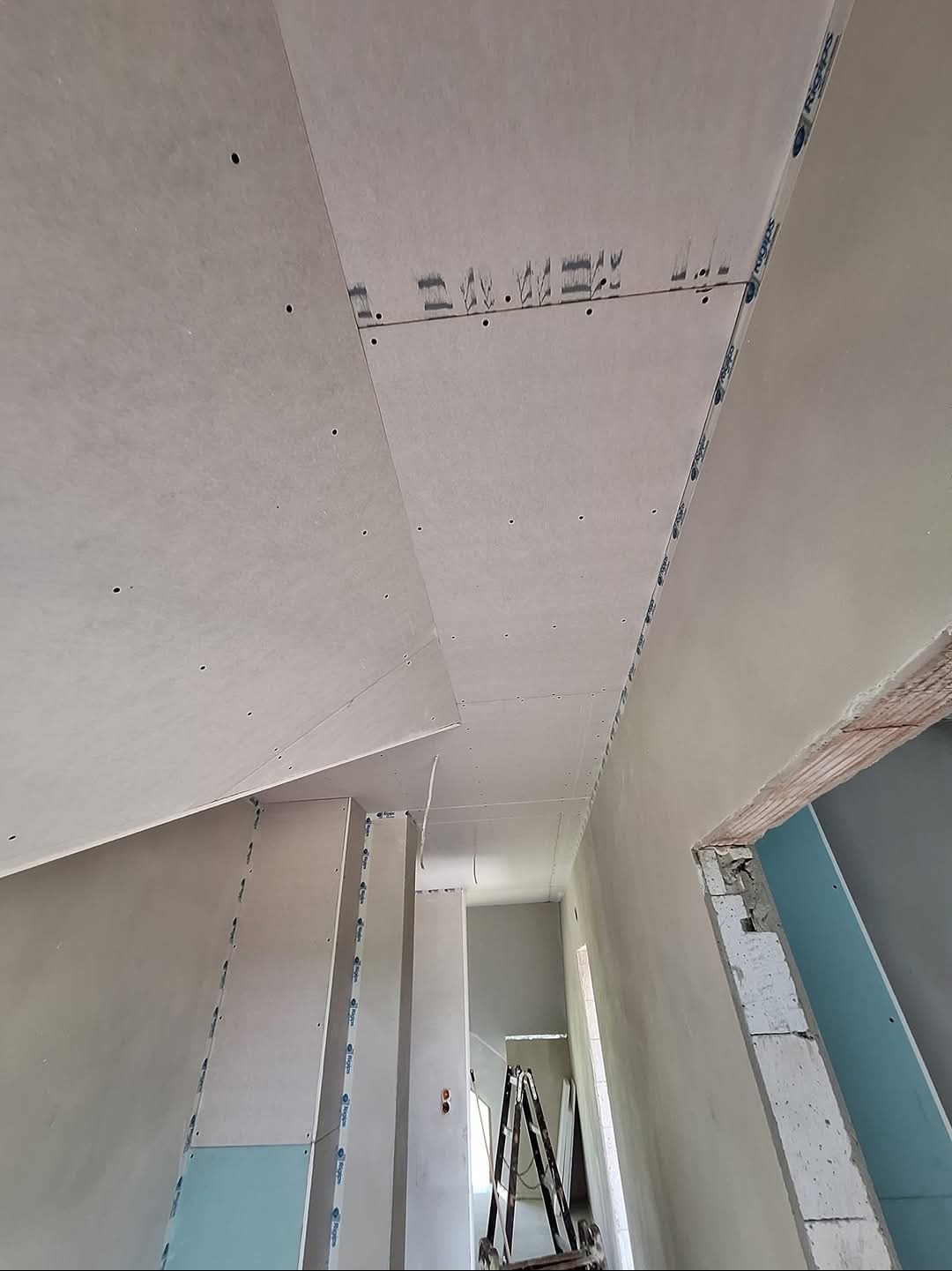 Montaż płyt kartonowo-gipsowych na suficie poddasza, widoczne łączenia płyt wzmocnione taśmą, surowe ściany i konstrukcja otworu drzwiowego, w tle drabina.