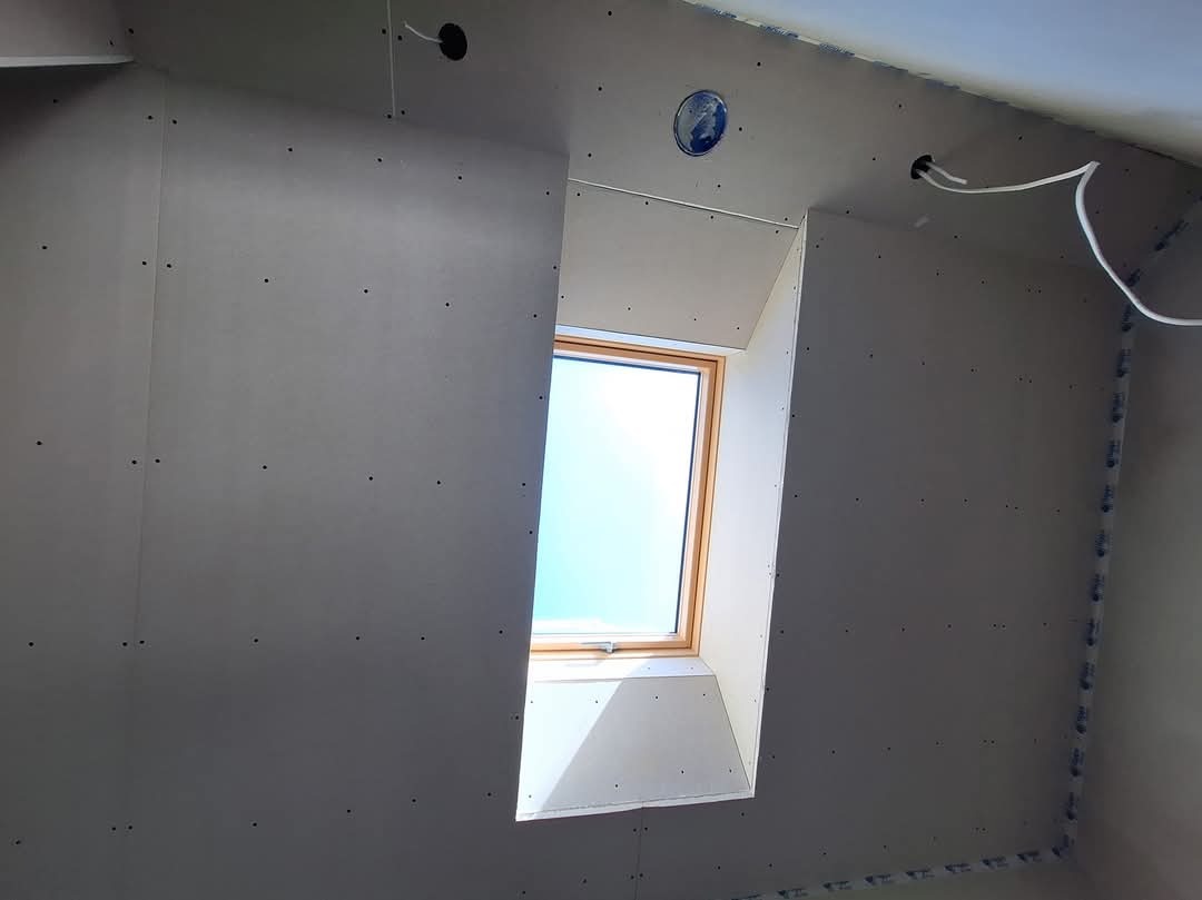 Montaż płyt kartonowo-gipsowych wokół okna dachowego, widoczne wkręty mocujące i wystające przewody elektryczne.