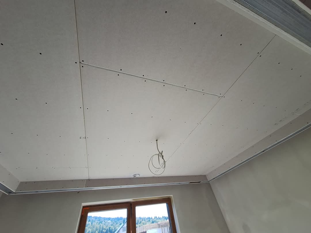 Montaż sufitu podwieszanego z płyt kartonowo-gipsowych, widoczne profile metalowe i instalacja elektryczna, okno w tle.