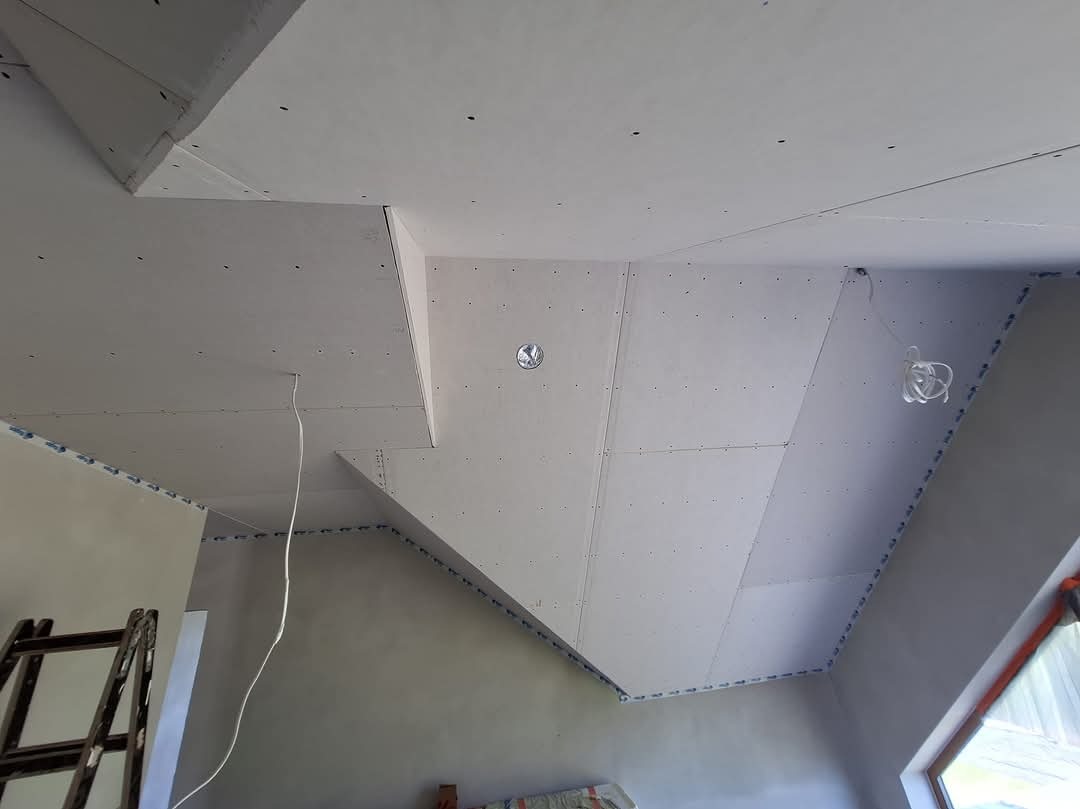 Skośny sufit poddasza z płyt kartonowo-gipsowych w trakcie montażu, widoczne łączenia płyt, przewody elektryczne i drabina oparta o ścianę.