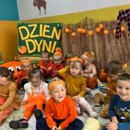Grupa uśmiechniętych dzieci w pomarańczowych ubraniach i opaskach z dyniami siedzi na sianie na tle dekoracji z okazji Dnia Dyni w żłobku.