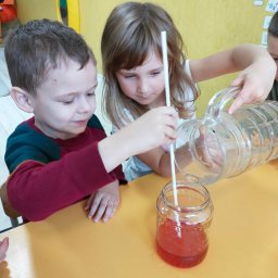 Przedszkole Niepubliczne Goleniów Wesołe Smyki - Dwoje dzieci przy żółtym stoliku w trakcie eksperymentu z przelewaniem czerwonego płynu z dzbanka do słoika, w którym znajduje się słomka.