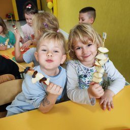 Przedszkole Niepubliczne Goleniów Wesołe Smyki - Dwoje uśmiechniętych dzieci trzymających szaszłyki owocowe z jabłek i śliwek przy żółtym stole, widoczne również inne dzieci w tle, na ramieniu chłopca widoczny tatuaż.