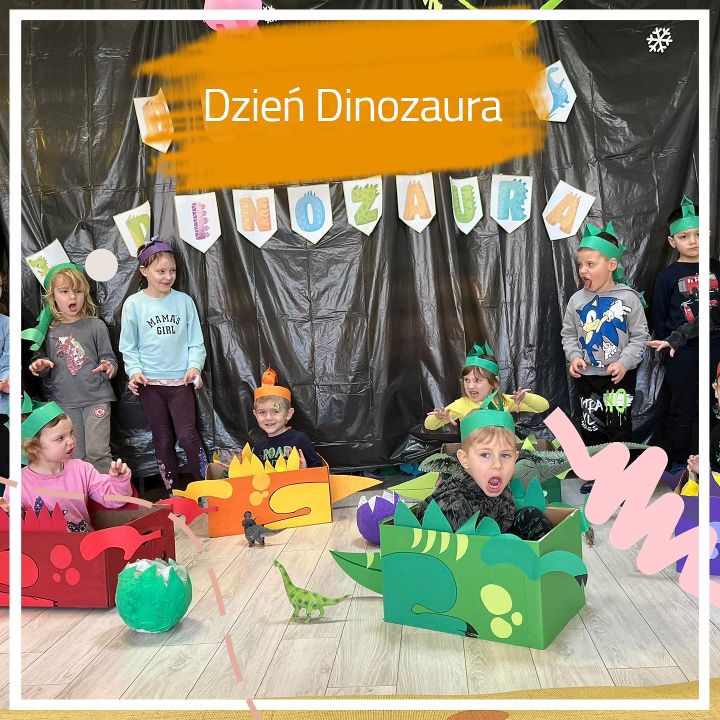 Grupa dzieci w kartonowych kostiumach dinozaurów świętuje 'Dzień Dinozaura', z dekoracjami i banerem w tle.