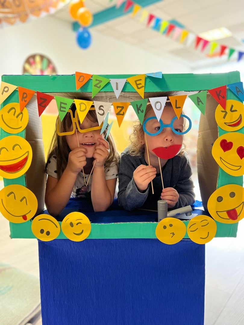 Dwie dziewczynki w tekturowej budce z napisem 'Przedszkola', udekorowanej emotikonami, trzymają rekwizyty do zdjęć: okulary i usta na patyku.