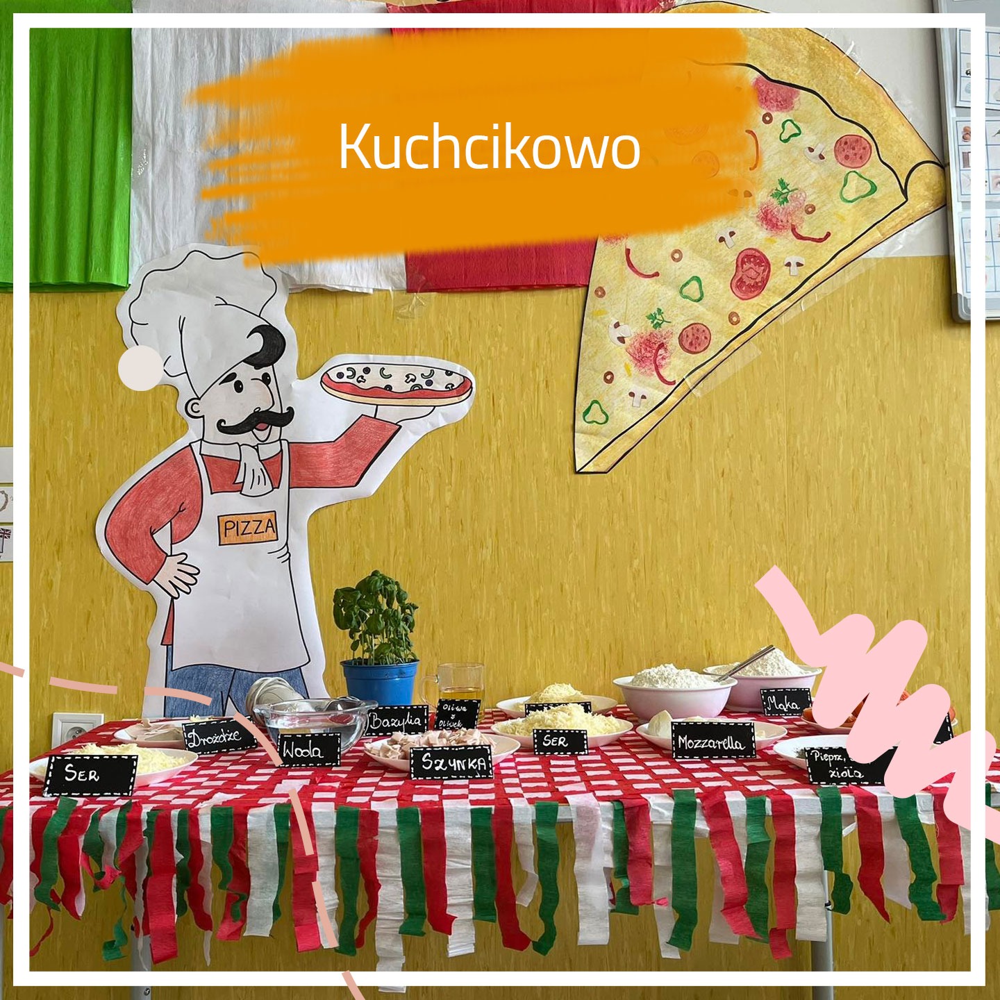 Dekoracja sali z motywem pizzy: kartonowy kucharz z pizzą, składniki na stole z etykietami, duży rysunek pizzy na ścianie, flaga Włoch w tle, napis Kuchcikowo.